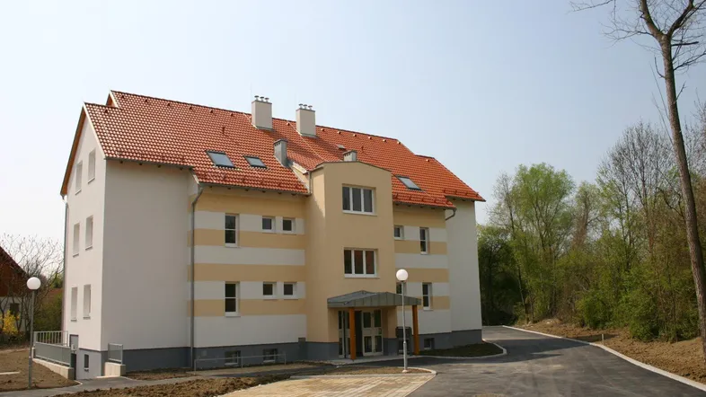 Neues Mehrfamilienhaus mit heller Fassade, rotem Ziegeldach und gepflastertem Eingangsbereich.
