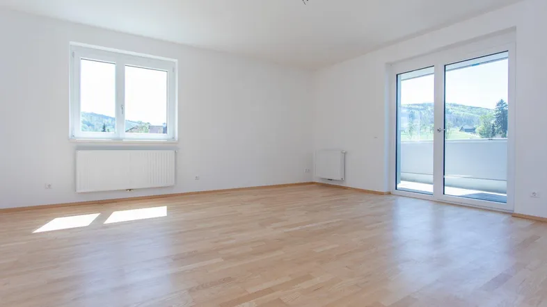 Helles, leeres Zimmer mit Parkettboden, großen Fenstern und Zugang zu einem Balkon.