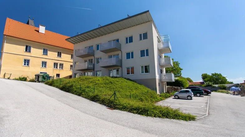 Modernes Mehrfamilienhaus mit Balkonen und Parkplätzen, umgeben von Grünflächen und einem historischen Gebäude.