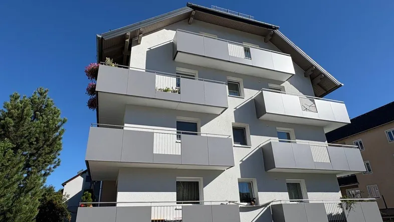 Modernes Mehrfamilienhaus mit mehreren grauen Balkonen und gepflegter Grünfläche unter strahlend blauem Himmel.