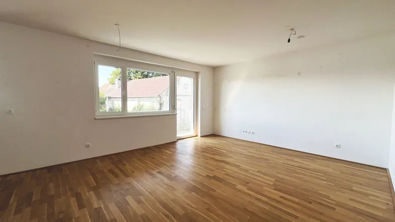 Helles, leeres Wohnzimmer mit Parkettboden und Zugang zu einem Balkon oder einer Terrasse.