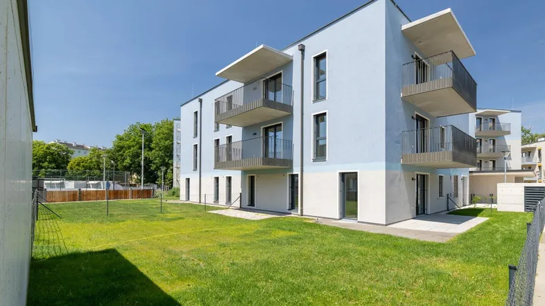 Modernes Mehrfamilienhaus mit hellblauer Fassade, mehreren Balkonen und einer weitläufigen Grünfläche. Im Hintergrund sind weitere Gebäude und Bäume sichtbar.
