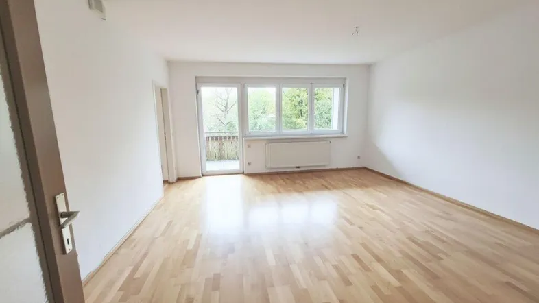 Helles Wohnzimmer mit Parkettboden, weißen Wänden und Zugang zu einem Balkon mit Blick ins Grüne.
