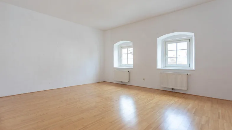 Helles, leeres Zimmer mit Parkettboden und zwei Fenstern, bereit zur individuellen Gestaltung.