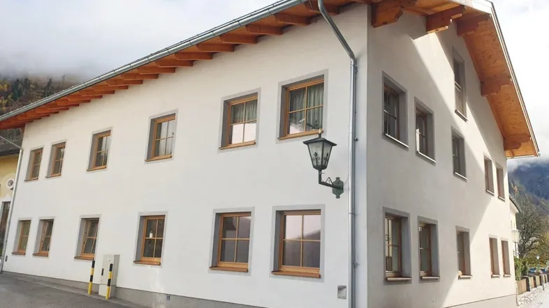 Traditionelles Gebäude mit weißer Fassade, Holzbalken und vielen Fenstern in alpiner Umgebung.