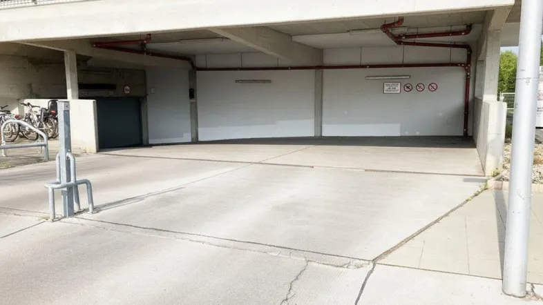 Eingangsbereich einer Tiefgarage mit Betonrampe und Fahrradstellplätzen im Freien, rote Rohre sind an der Decke sichtbar.