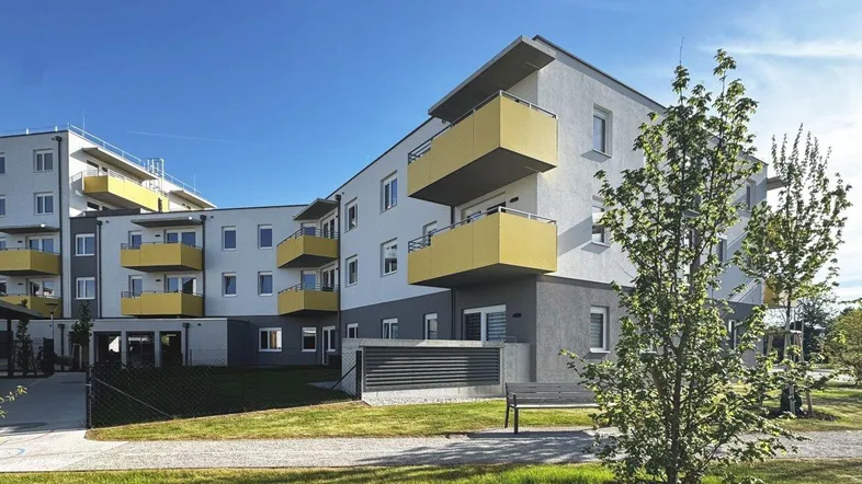 Modernes Mehrfamilienhaus mit gelben Balkonen und gepflegter Grünanlage unter blauem Himmel.