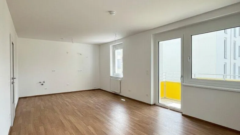 Helles, leeres Wohnzimmer mit Laminatboden und Zugang zu einem Balkon.