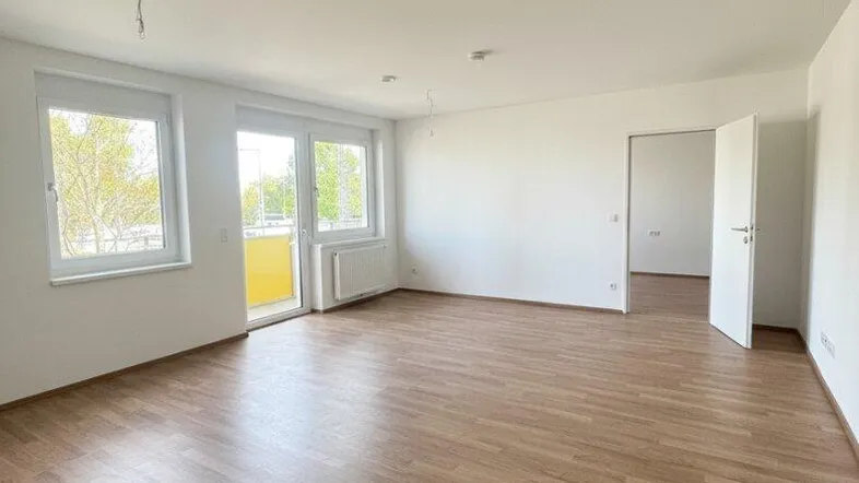 Helles, leeres Zimmer mit Laminatboden, weißen Wänden und Zugang zu einem Balkon.