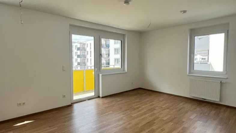Helles, leeres Wohnzimmer mit Laminatboden und Zugang zu einem Balkon.