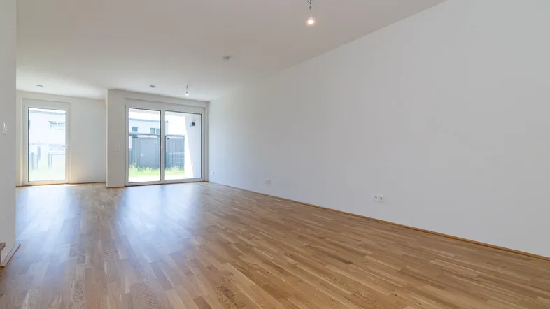 Heller, leerer Wohnbereich mit Parkettboden und Zugang zur Terrasse durch große Schiebetüren.