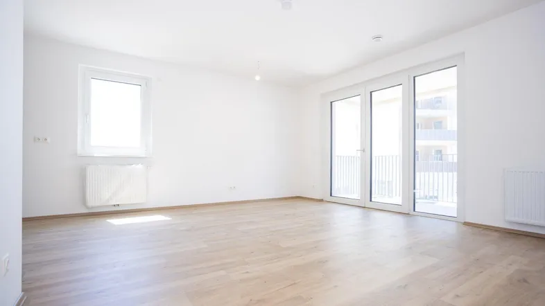 Helles, leeres Zimmer mit Laminatboden und Zugang zu einem Balkon, ideal für Wohn- oder Schlafbereich.