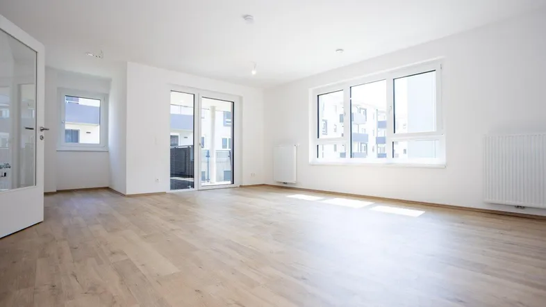 Helles, leeres Wohnzimmer mit großen Fenstern, Laminatboden und Zugang zum Balkon.