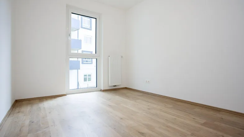 Helles Zimmer mit Laminatboden, weißen Wänden und einem Fenster mit Blick auf andere Gebäude.