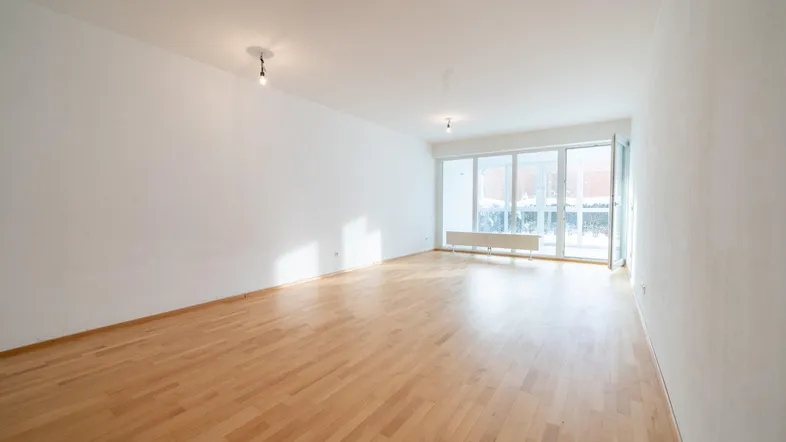 Helles, leeres Wohnzimmer mit Parkettboden und großen Fenstern, die viel Tageslicht hereinlassen.