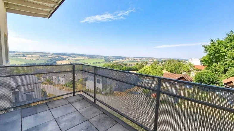 Geräumiger Balkon mit Fliesenboden und weitem Blick über die grüne Landschaft und umliegende Häuser.