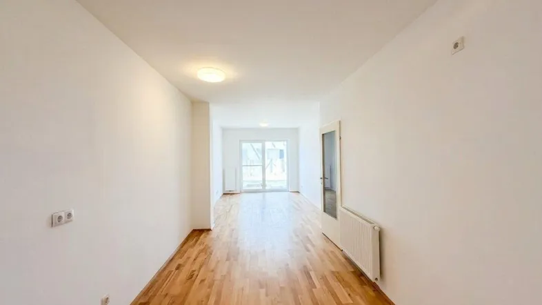 Helles, leeres Zimmer mit Parkettboden und Zugang zu einem Balkon oder einer Terrasse.