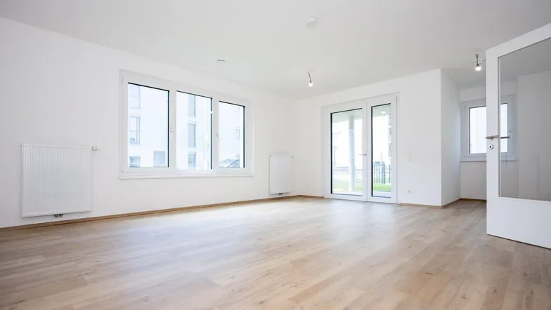 Helles, leeres Wohnzimmer mit Laminatboden, großen Fenstern und Zugang zur Terrasse.