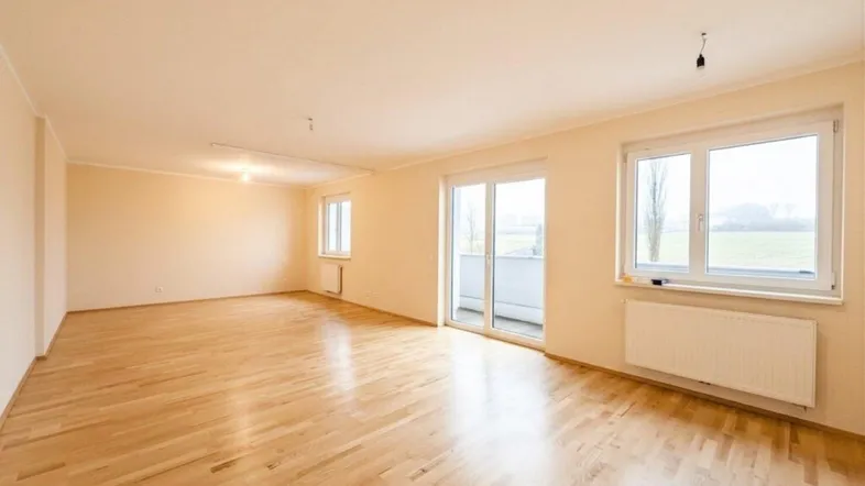 Heller, leerer Raum mit Holzfußboden, großen Fenstern und Zugang zu einem Balkon mit grüner Aussicht.