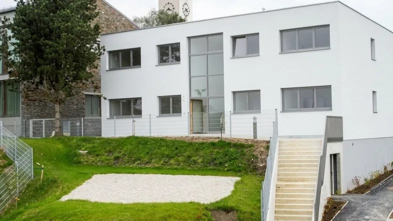Modernes weißes Gebäude mit großen Fenstern, umgeben von einer grünen Landschaft und einer Kirche im Hintergrund.