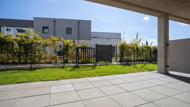 Große gepflasterte Terrasse mit Rasenfläche und Blick auf moderne Wohngebäude unter blauem Himmel.