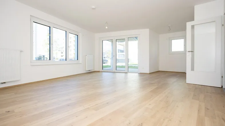 Helles, leeres Wohnzimmer mit großen Fenstern, hellem Laminatboden und Zugang zur Terrasse.
