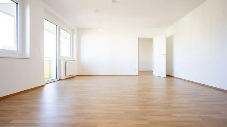 Helles, leeres Zimmer mit Laminatboden und Zugang zu einem Balkon.