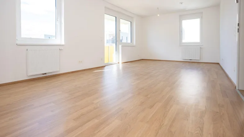 Helles, leeres Zimmer mit Laminatboden, weißen Wänden und Zugang zu einem Balkon.