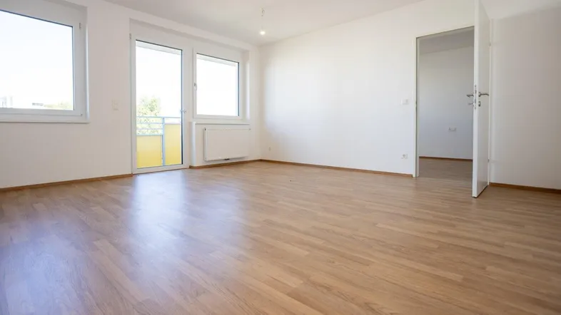 Heller Wohnraum mit Laminatboden, weißen Wänden und Zugang zu einem Balkon durch eine große Tür.