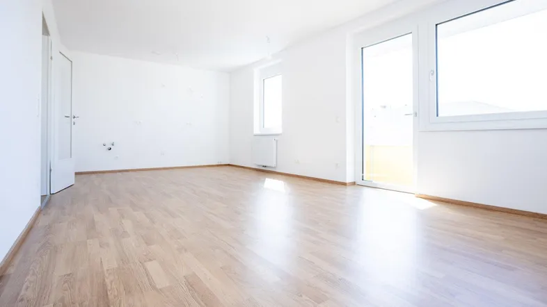 Helles, leeres Zimmer mit Laminatboden, weißen Wänden und Zugang zu einem Balkon.