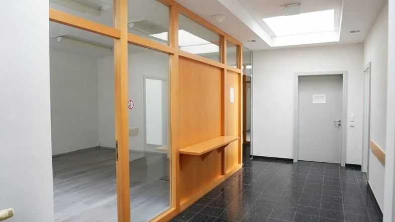 Heller Flur mit Holz-Glas-Trennwänden und dunklem Fliesenboden, der zu Büroräumen führt.