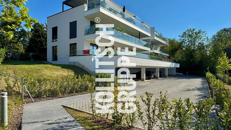 Modernes Mehrfamilienhaus mit Balkonen, Glasgeländern und Tiefgaragenzufahrt in sonniger Umgebung.
