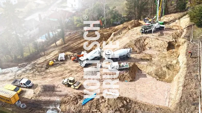 Luftaufnahme einer Baustelle am Hang mit mehreren Baufahrzeugen und Aushubarbeiten für ein neues Gebäude.