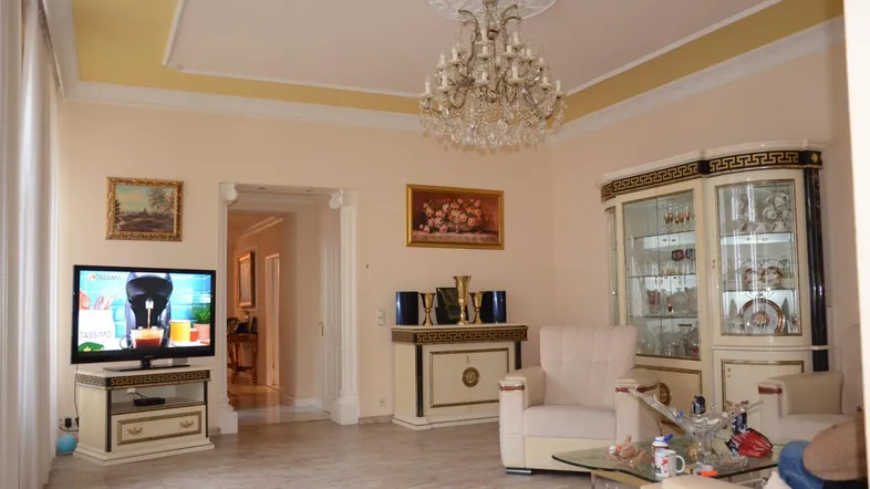 Geräumiges Wohnzimmer mit klassischem Kronleuchter, Fliesenboden und eleganten Möbeln in hellem Ambiente.