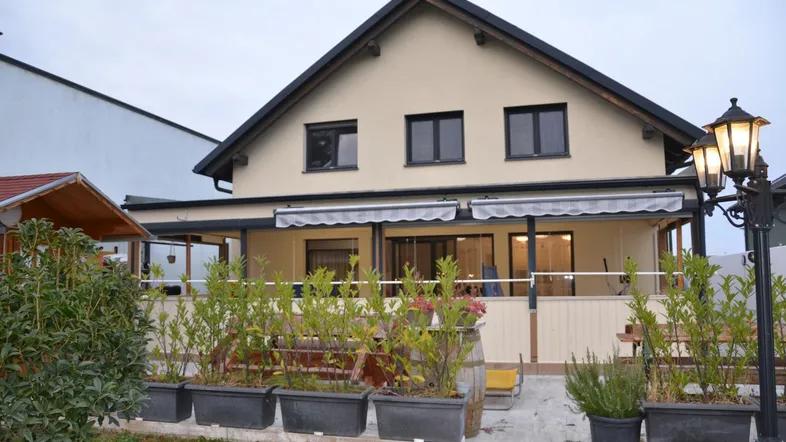 Hausfassade mit überdachter Terrasse, bepflanzten Blumenkästen und Gartenbeleuchtung am Abend.