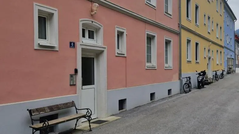 Mehrere farbige Gebäude entlang einer Straße unter bewölktem Himmel, mit einer Bank vor dem rosafarbenen Haus.