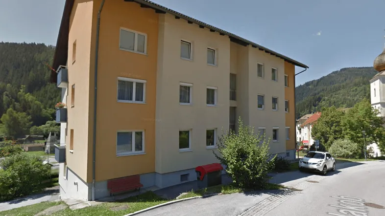 Mehrfamilienhaus mit schräger Dachkonstruktion, orangefarbener Fassade und umliegender Naturkulisse.