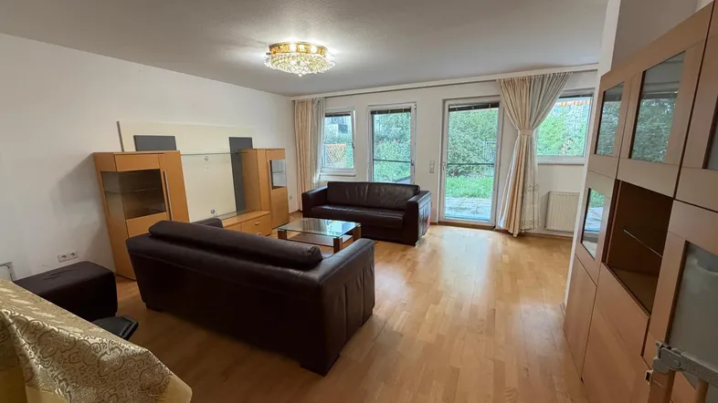 Helles Wohnzimmer mit zwei Ledersofas, Holzmöbeln und Zugang zu einem grünen Außenbereich.