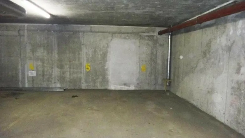 Tiefgaragenstellplatz mit Betonwänden und -boden, beleuchtet mit Leuchtstoffröhren und sichtbaren Rohren an der Decke.