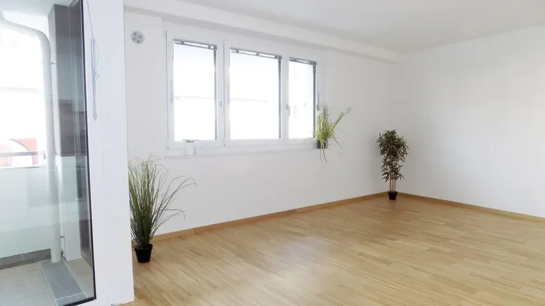 Helles, leeres Zimmer mit Parkettboden, großen Fenstern und Zugang zu einem Balkon.