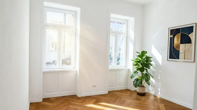 Helles Zimmer mit Fischgrätparkettboden und zwei großen Fenstern, das viel Tageslicht hereinlässt.