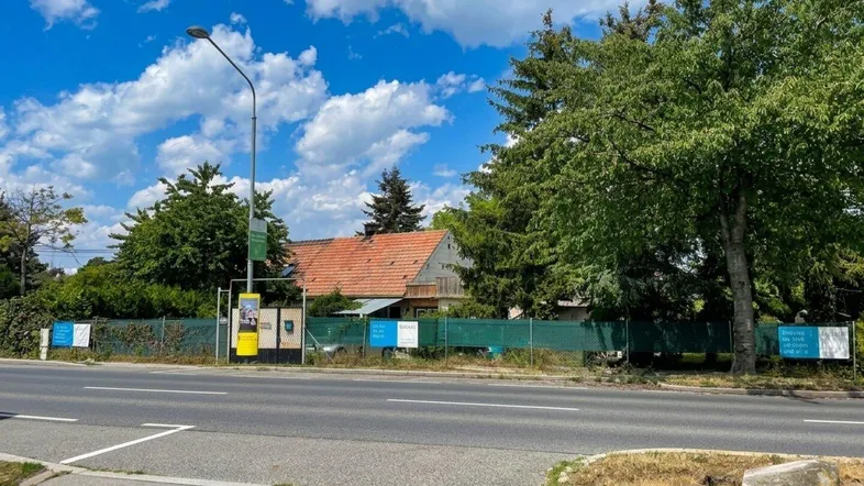 Blick von der Straße auf ein Grundstück mit altem Haus, umgeben von Bäumen und einem Zaun unter blauem Himmel.