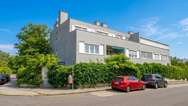 Modernes Mehrfamilienhaus mit grauer Fassade und begrüntem Vorgarten an der Straße.