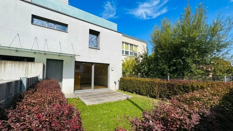 Blick auf die Rückseite des modernen Gebäudes mit einer kleinen Terrasse und einem gepflegten Gartenbereich.