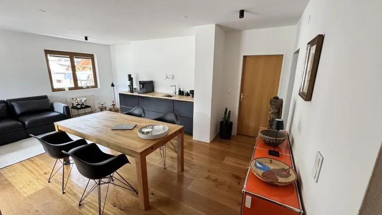 Heller offener Wohn- und Essbereich mit Holzboden, schwarzem Sofa und integrierter Küchenzeile.