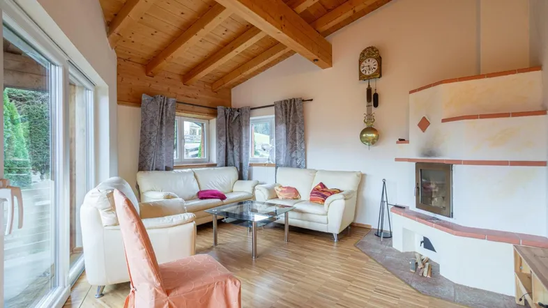 Gemütliches Wohnzimmer mit Holzbalkendecke, Kamin und großen Fenstern, die viel Licht hereinlassen.