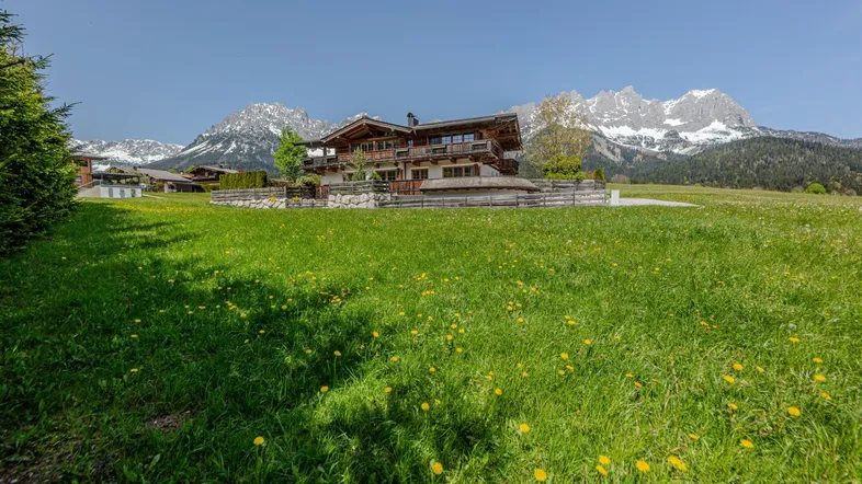 Großes Holzchalet in einer grünen Wiese mit Alpenpanorama und schneebedeckten Bergen im Hintergrund.