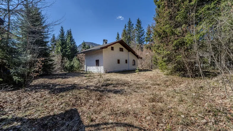 Freistehendes Haus in ländlicher Umgebung mit Wald und Bergen im Hintergrund unter blauem Himmel.