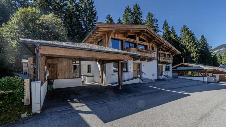 Modernes Chalet im alpinen Stil mit Holzfassade und integriertem Carport, umgeben von grünen Bäumen.