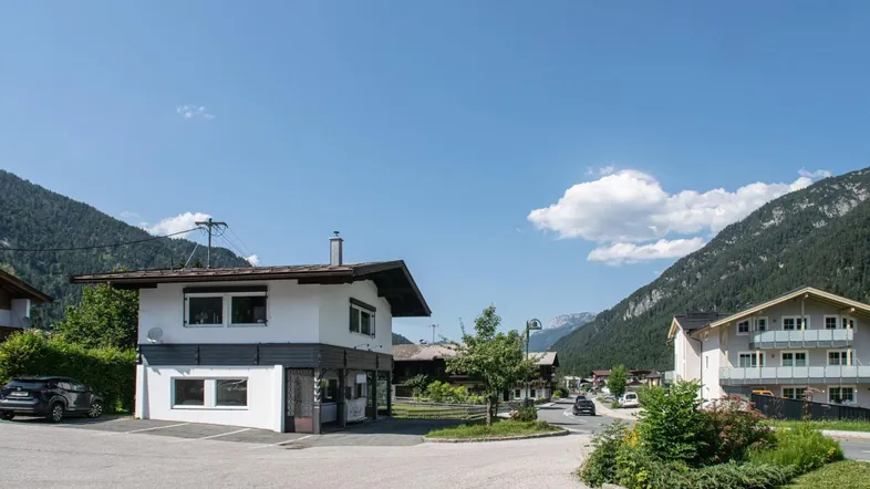 Ein weißes Gebäude mit dunklem Dach und Parkplätzen, umgeben von Bergen und grüner Landschaft.